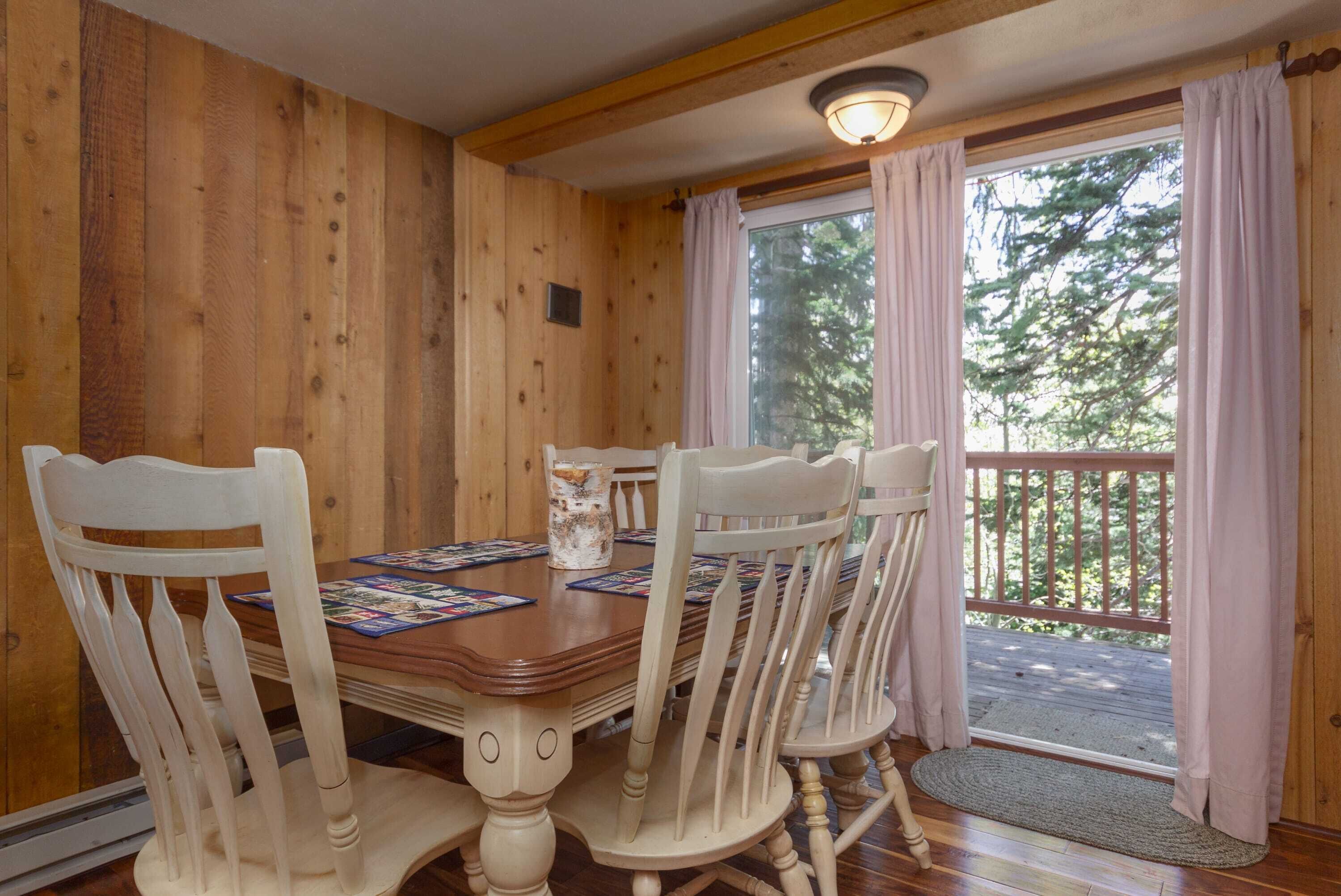 Interior de madera con una mesa de comedor, sillas, una ventana con cortinas y vistas al exterior.