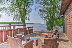 Een terras met rieten meubels met uitzicht op een meer, een houten omheining en een gebouw met een raam.