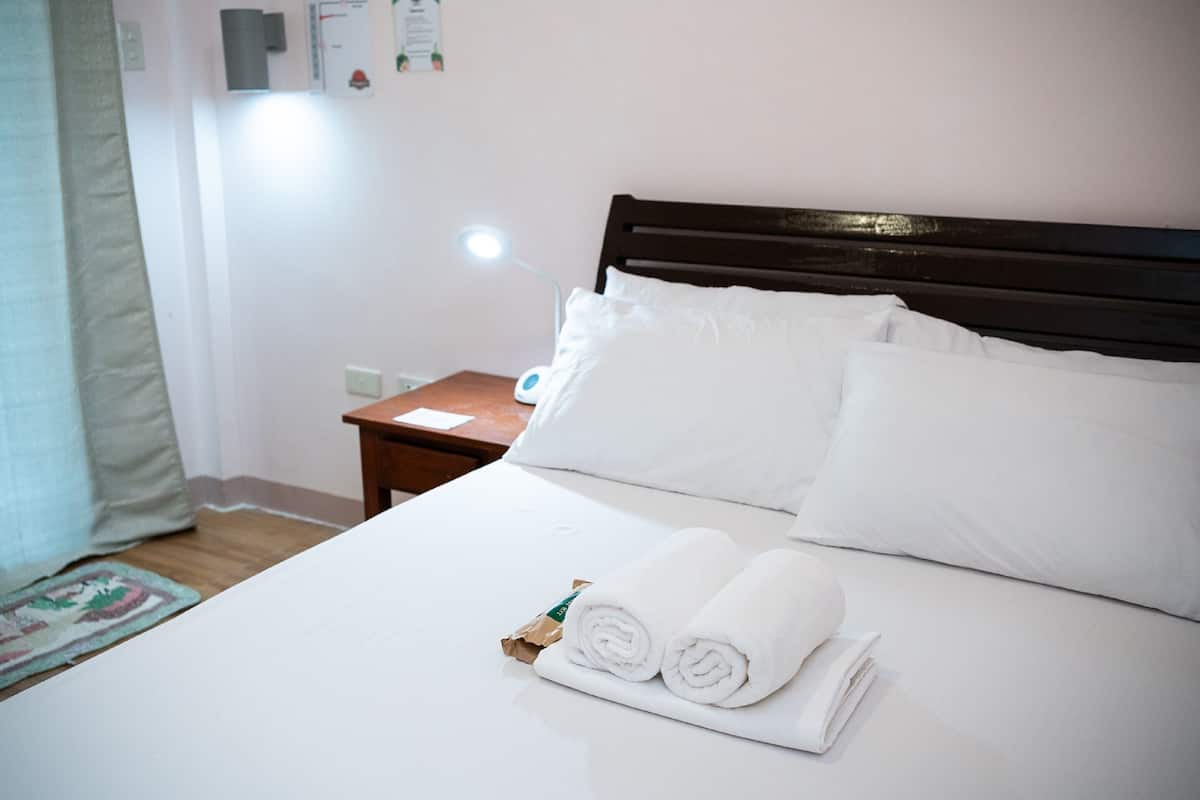 Un lit bien fait, avec du linge de lit blanc, une tête de lit en bois et une petite table de chevet sur laquelle se trouve une lampe.