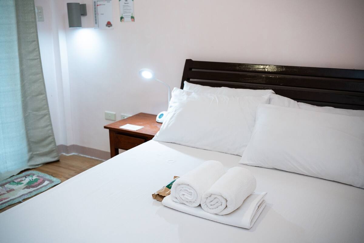 Un lit bien fait, avec du linge de lit blanc, une tête de lit en bois et une petite table de chevet sur laquelle se trouve une lampe.