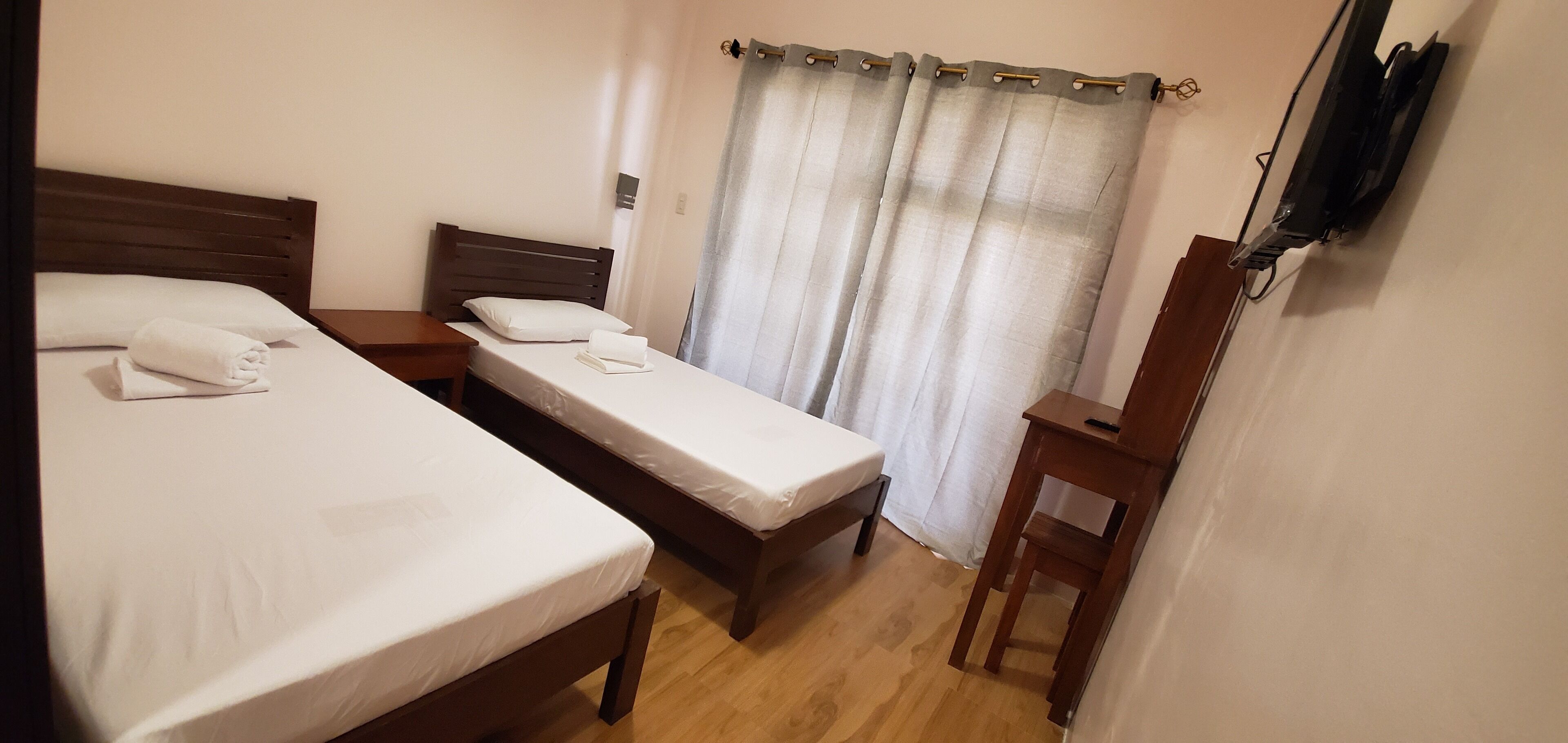 A room with two beds, a wooden nightstand, a TV mounted on the wall, and a curtain.