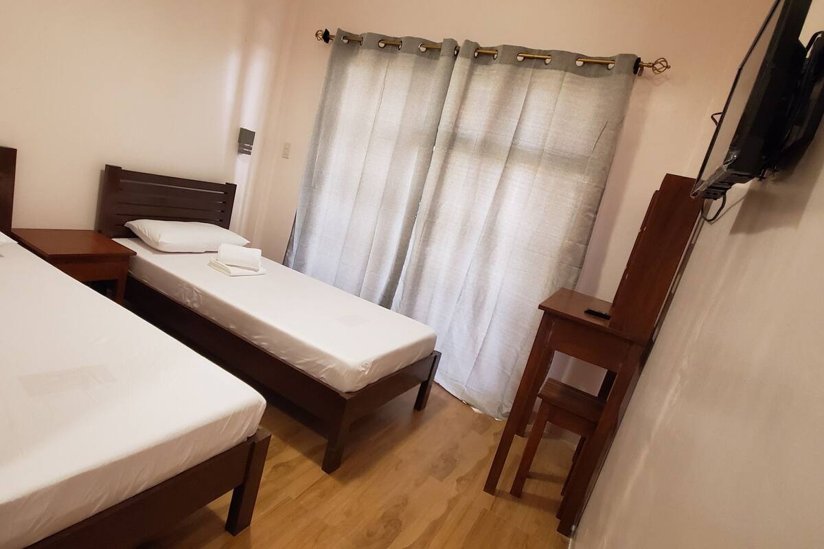 Une chambre avec deux lits, une table de chevet en bois, un téléviseur fixé au mur et un rideau.