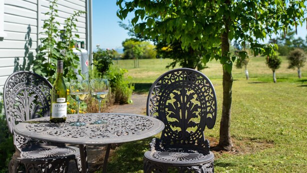 Une table ronde en métal, accompagnée de deux chaises, une bouteille de vin et deux verres, est placée à l’extérieur, avec une maison et des arbres en arrière-plan.