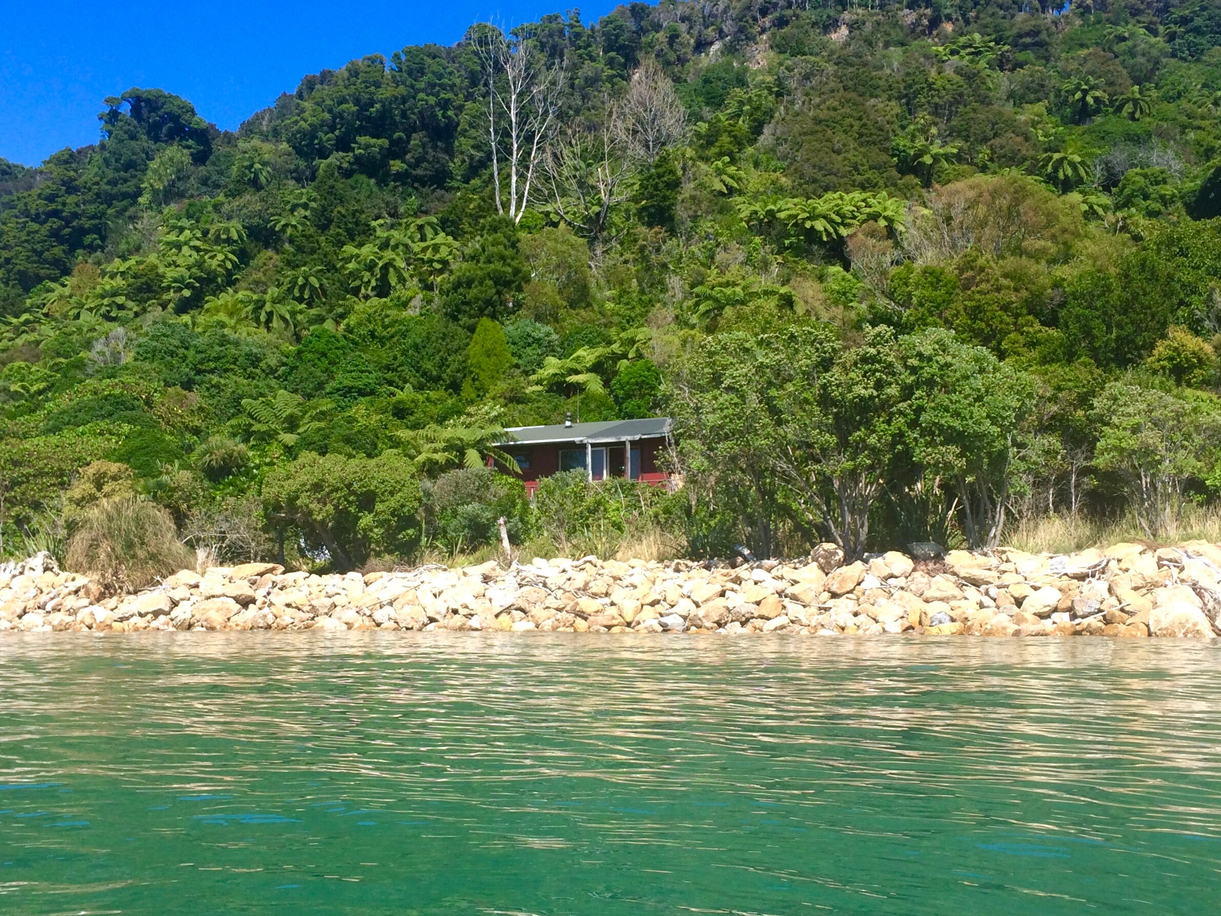 Una casa con techo rojo y balcón, rodeada por un bosque frondoso y un litoral rocoso.