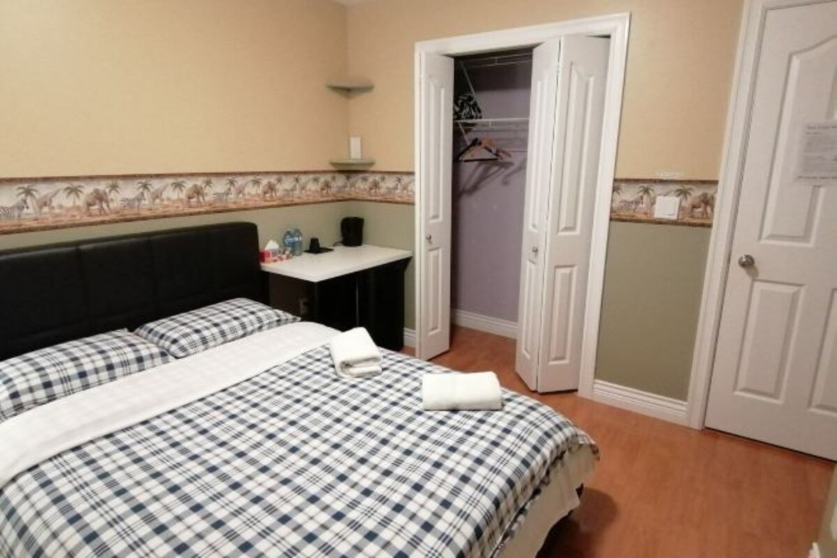 A neatly made bed with a checkered bedspread, a bedside table with a lamp, and a door leading to a closet with hanging clothes.