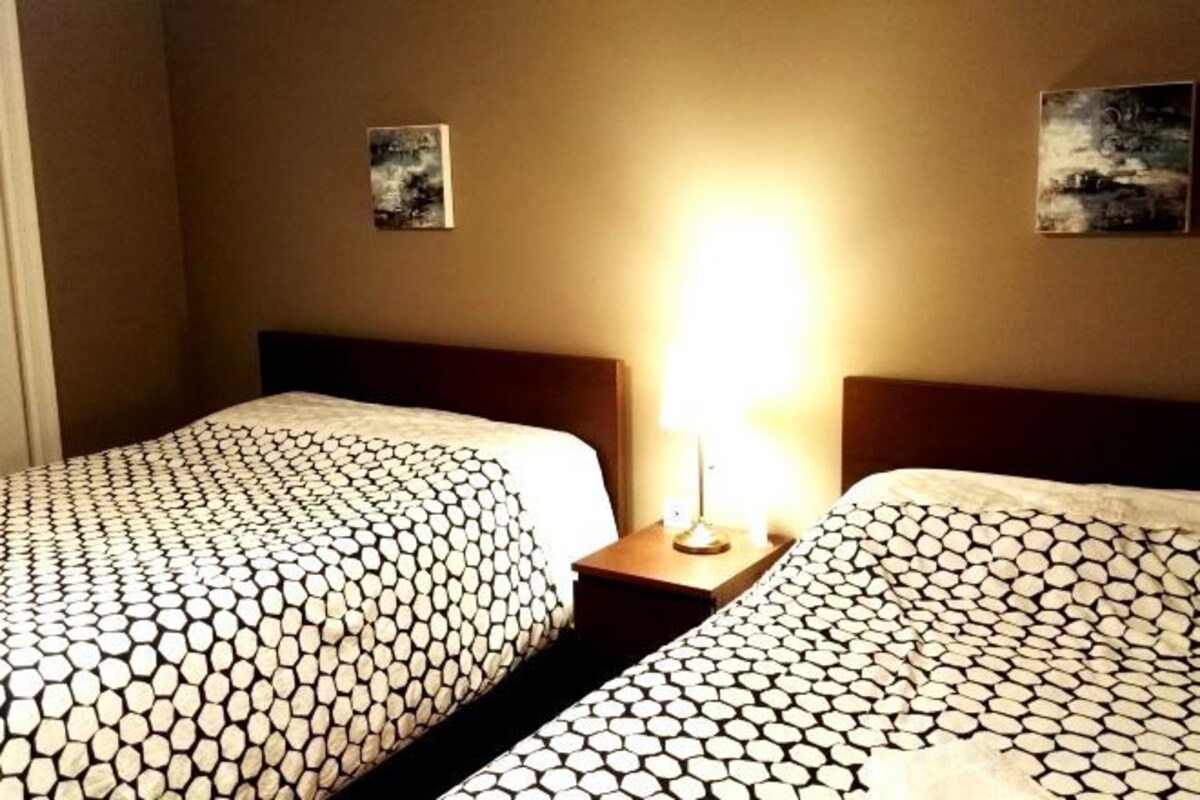 A hotel room with two beds, a nightstand with a lamp, and artwork on the walls.