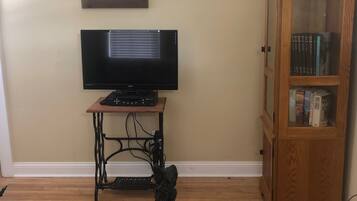 A living room with a television on a wooden stand, a sewing machine, a cabinet with books, and a small table with a plate and pinecones.