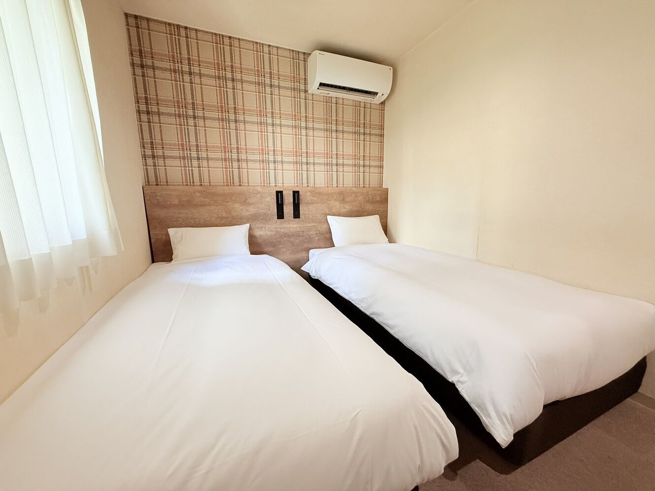 A double bed room with a plaid headboard, an air conditioning unit, and a window with sheer curtains.