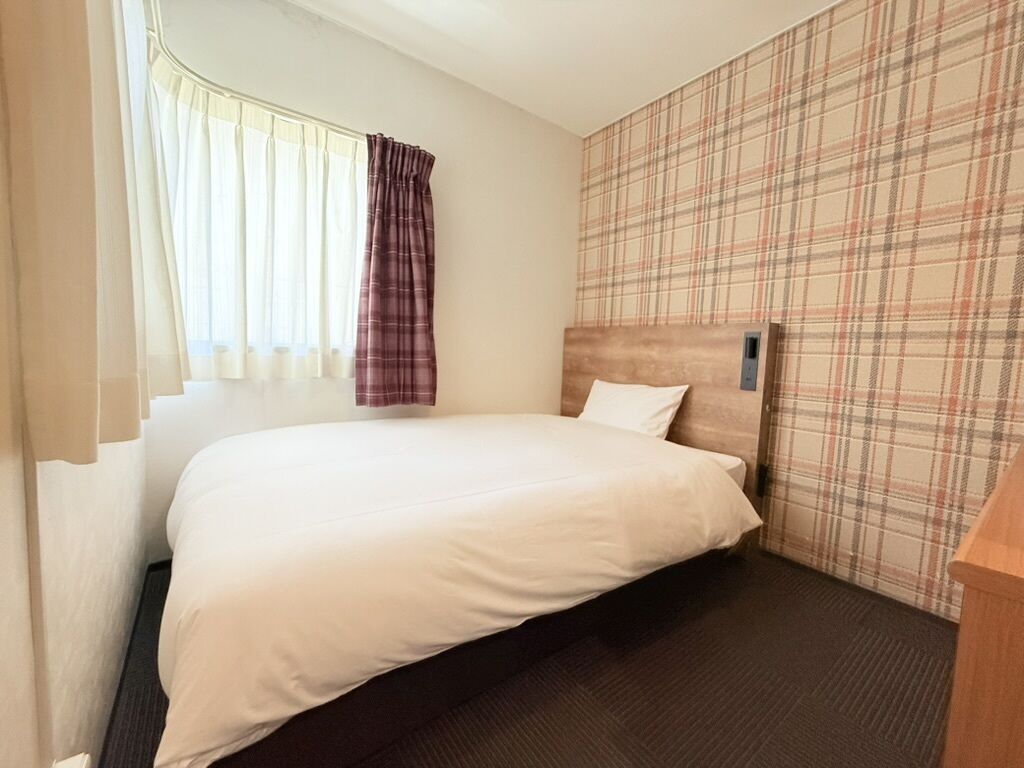 A hotel room with a bed, curtains, a plaid-patterned wall, and a wooden headboard.