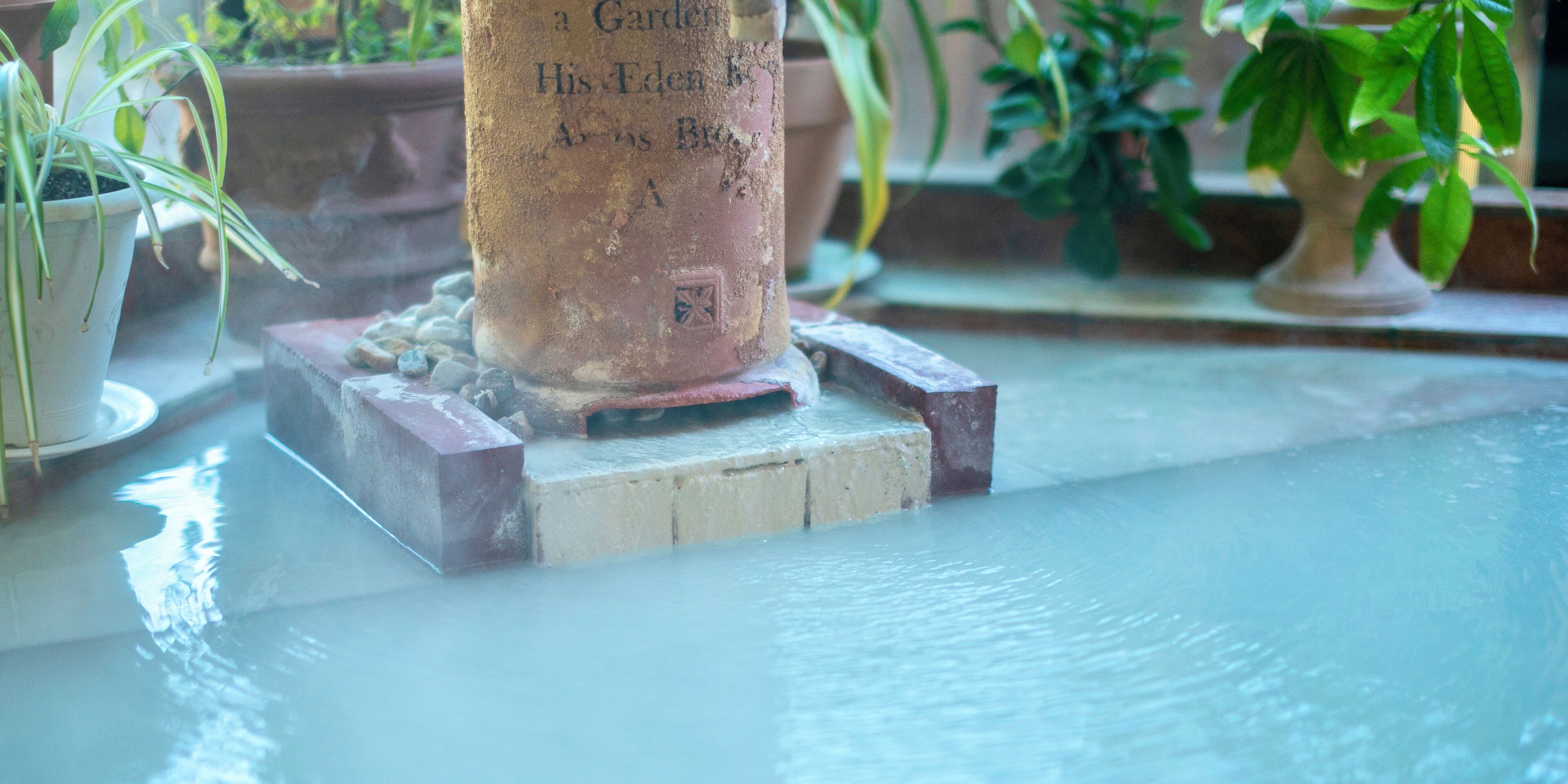 A water feature with a stone pedestal and a rectangular base, surrounded by potted plants.