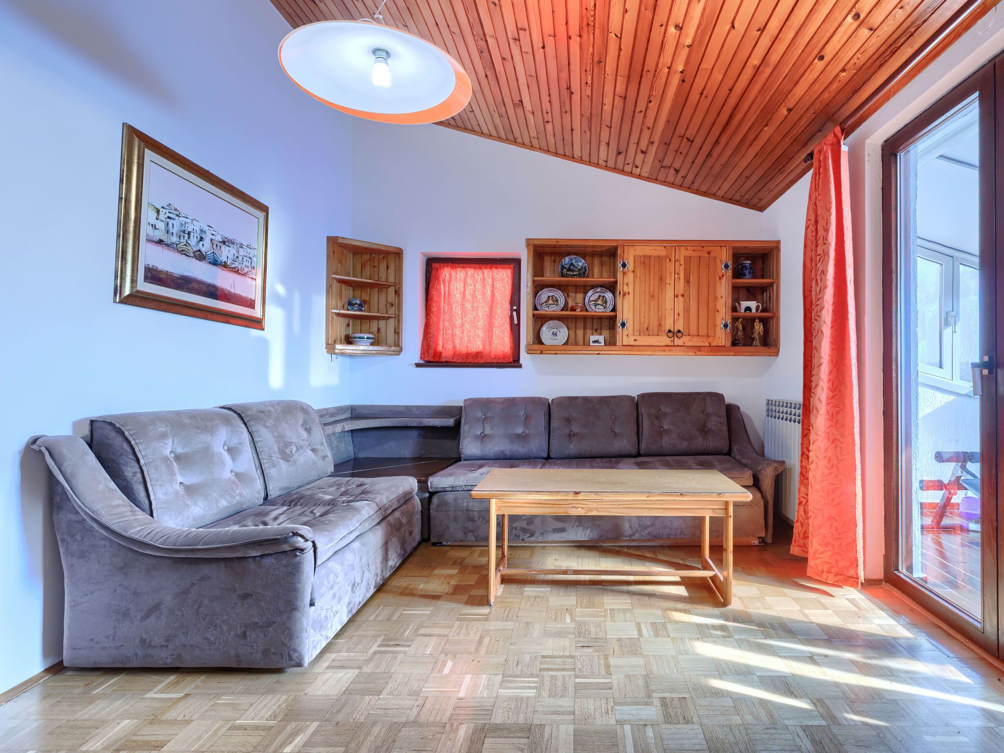 A living room with a grey sectional sofa, a wooden coffee table, and a herringbone-patterned floor. There is a wooden cabinet with dishes and a framed picture on the wall.