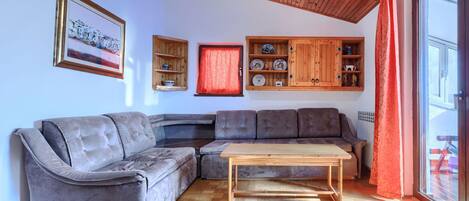 A living room with a grey sectional sofa, a wooden coffee table, and a herringbone-patterned floor. There is a wooden cabinet with dishes and a framed picture on the wall.