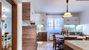 A modern kitchen with wooden cabinets, a dining area with a table and chairs, and a window with lace curtains. - Eufemia by Interhome (Pula)