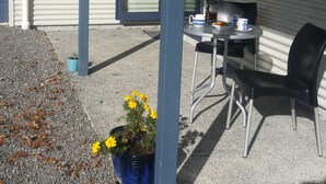 Un espace extérieur abrité avec une table et des chaises, un pot de fleurs contenant des fleurs jaunes et un bâtiment dont la façade est recouverte de tôle ondulée.