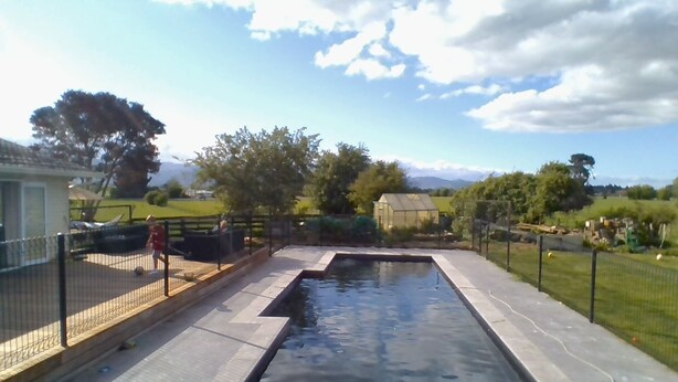 Une piscine dotée d’une terrasse en bois et d’une clôture en métal, entourée de verdure et surplombée par un ciel dégagé.