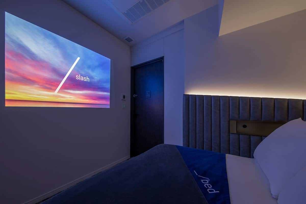 A hotel room with a bed, a headboard, a door, and a wall-mounted screen displaying a sunset scene.