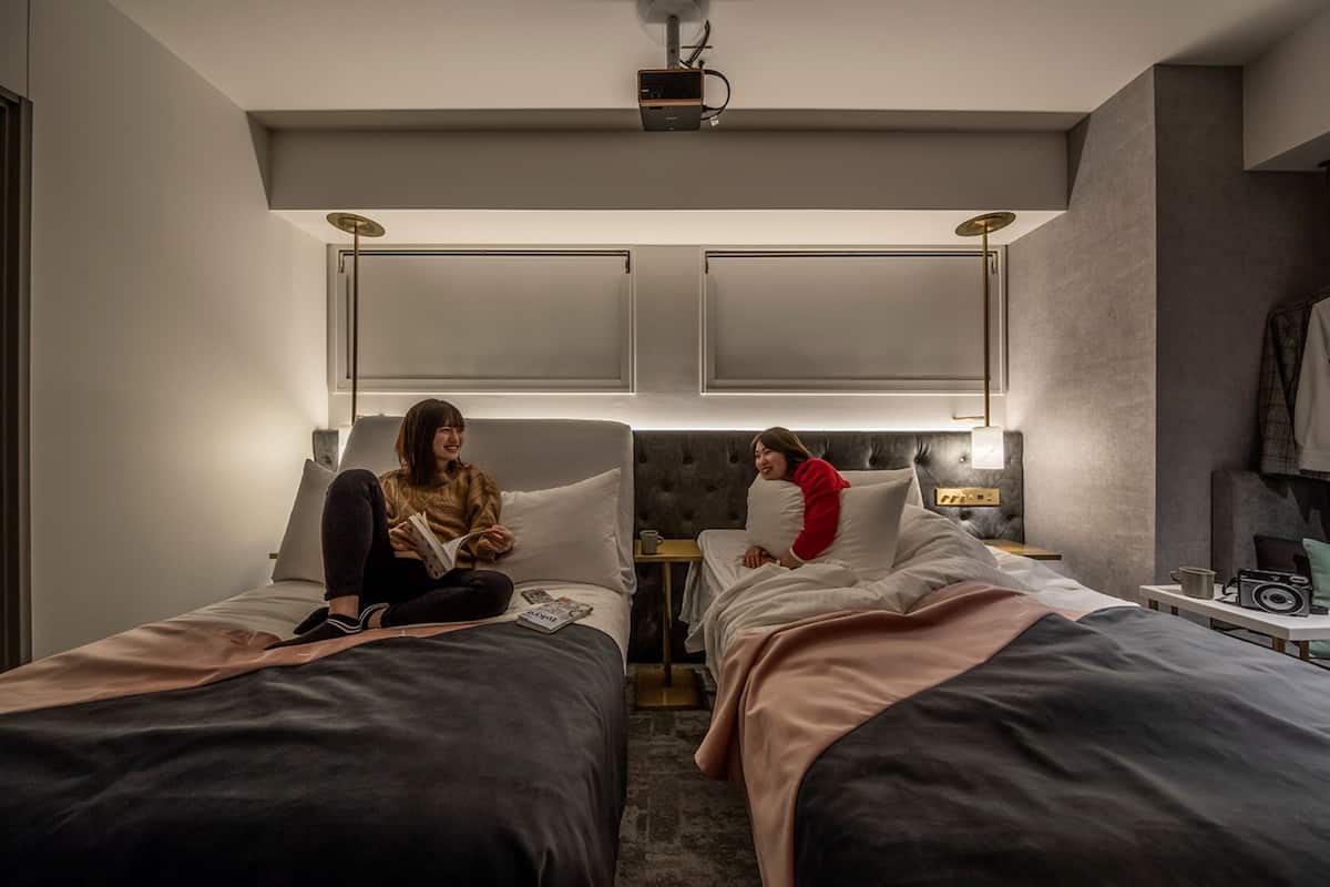 A hotel room with two beds, a ceiling projector, and a small table with a cup and a radio.