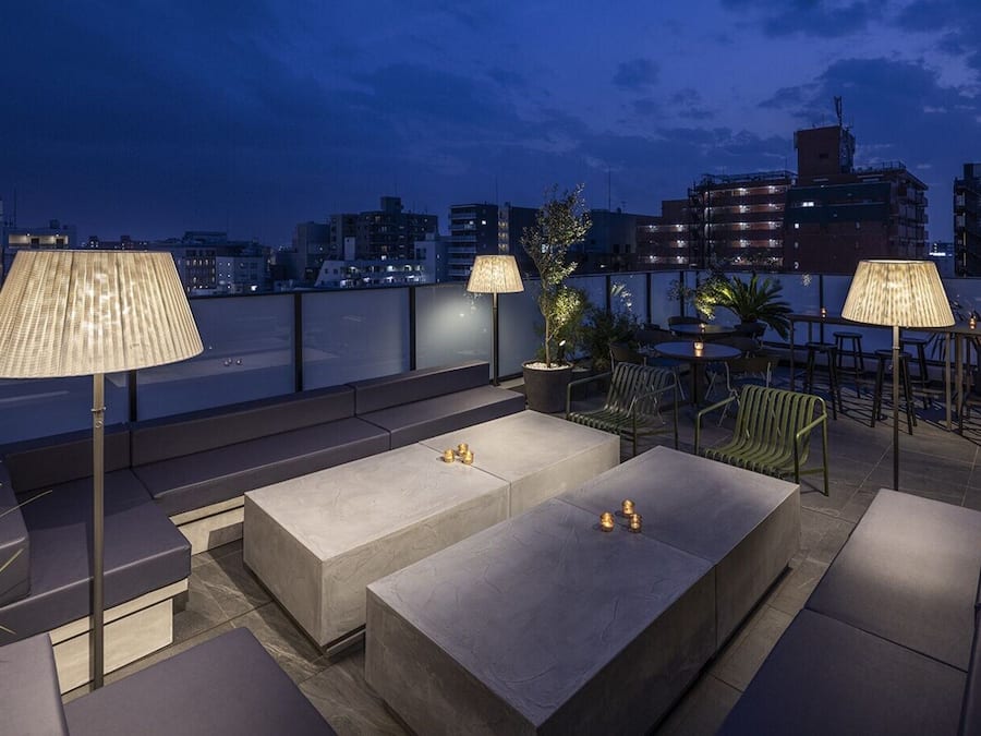 Un área de estar al aire libre con muebles y lámparas modernos, con vista a un paisaje urbano al atardecer.