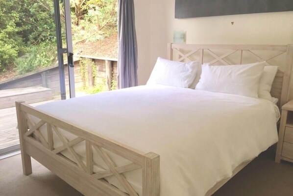 A bedroom with a wooden bed frame, white bedding, and a view of greenery outside. - Waiheke Oneroa - Luxury 3 bed room 2 bath room home with sweeping sea views (Oneroa)