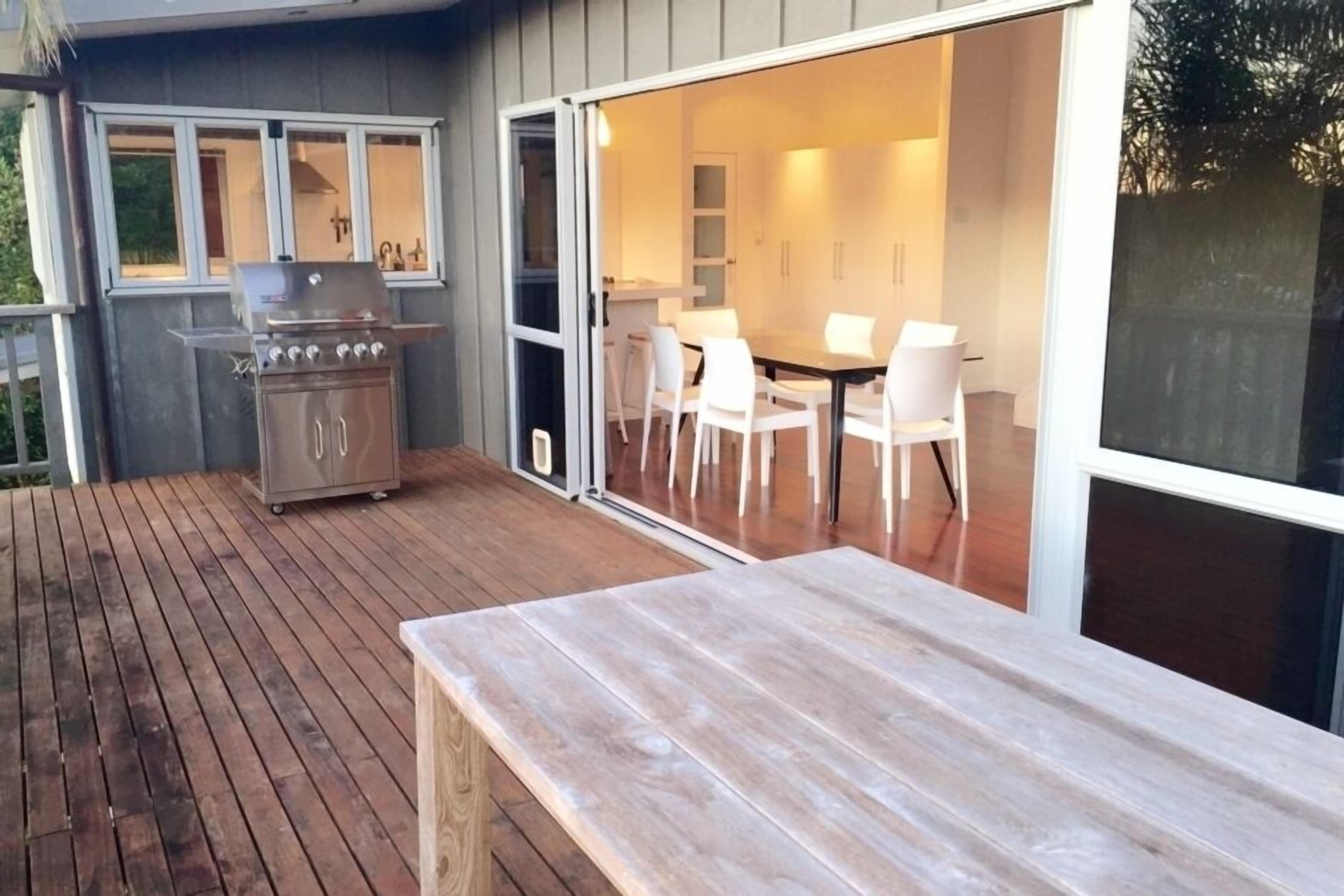 Une terrasse extérieure dotée d’une plateforme en bois, d’un barbecue et d’un coin repas avec des chaises et une table blanches.