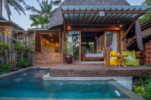 Un espace aménagé au bord de la piscine, comprenant un coin salon couvert, une clôture en bois et des plantes tropicales.