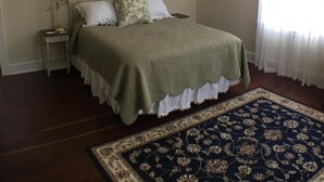 A bedroom with a bed, a ceiling fan, a nightstand, a lamp, a framed picture, and a window with curtains. - 2 Melinda’s Garden Apt  (Natchez)