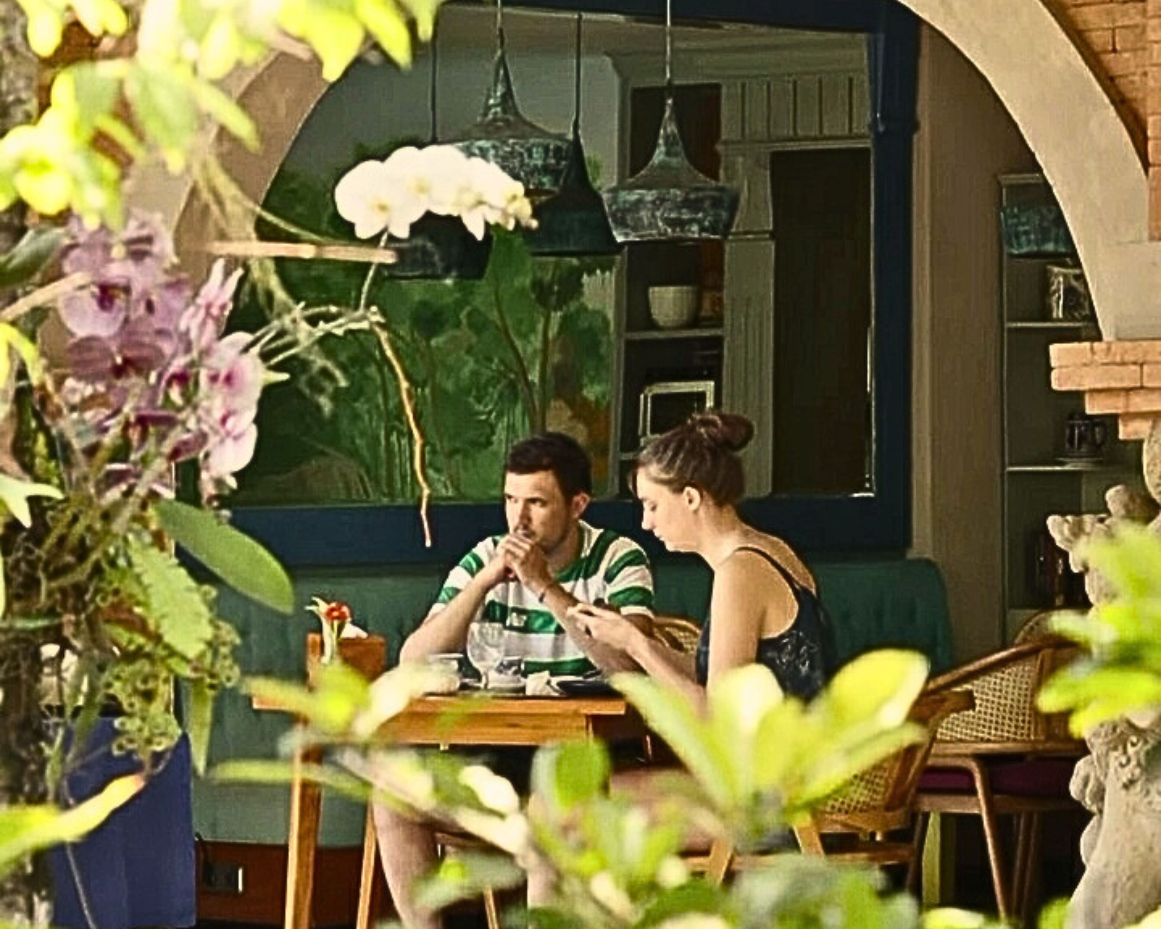Una pareja sentada en una mesa en un ambiente exterior acogedor, con un muro de ladrillo y follaje al fondo.