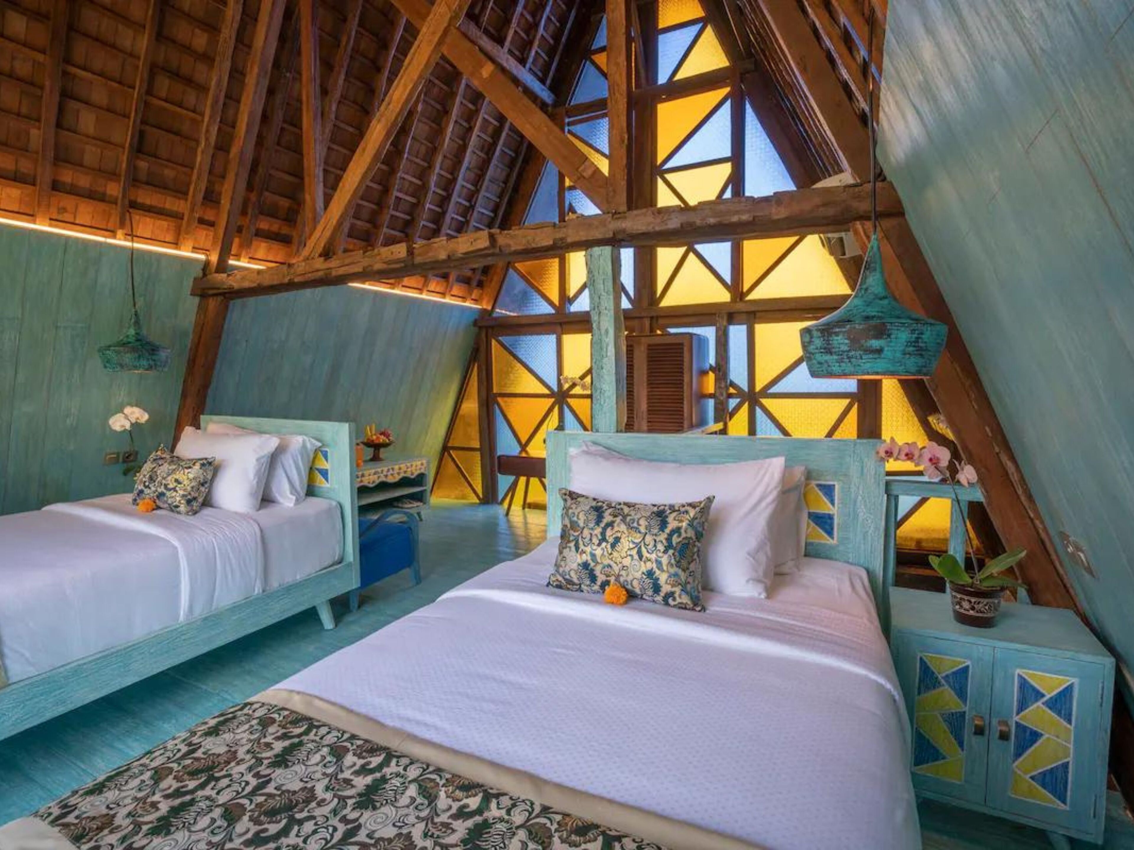 A cozy attic bedroom with two beds, a hanging light fixture, and a view through a geometric window.