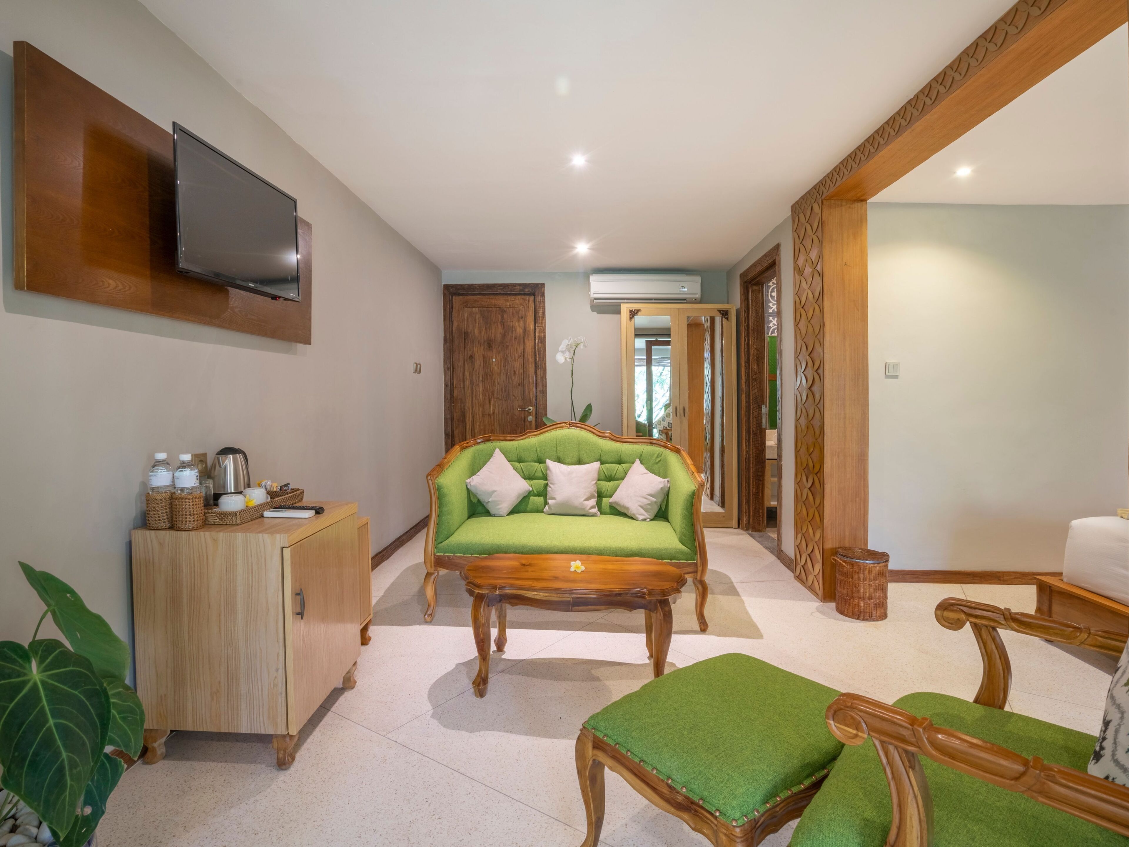 A spacious room with a green sofa, wooden furniture, a TV mounted on the wall, and a large mirror.