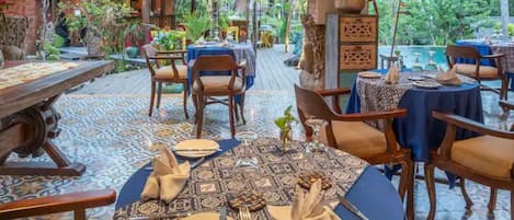 An outdoor dining area with tables set for two, surrounded by tropical plants and a pool in the background.