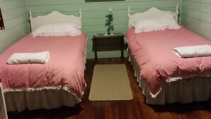 A room with two single beds, each with a pink bedspread and white pillows, a small wooden nightstand with a vase of flowers, and a beige rug on the wooden floor. - Te Whare Rural Retreat and Caravan (Havelock North)