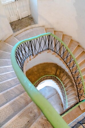 Staircase with a worn, spiral design, featuring a green handrail and black balusters, surrounded by white walls and a small window.
