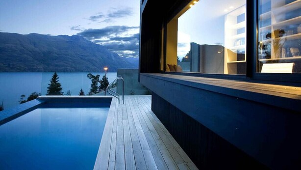 A modern building with a wooden deck and a pool overlooking a lake with mountains in the background.