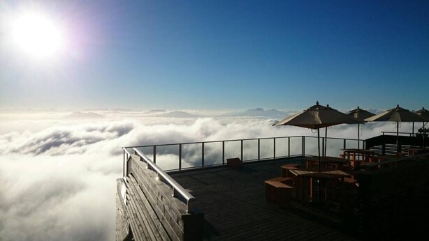 Una terraza de madera con mobiliario exterior y sombrillas, con vista a un mar de nubes.