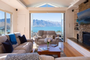 Relax in this serene living space with a panoramic view of the mountains and water, featuring a comfortable seating area and a cozy fireplace.