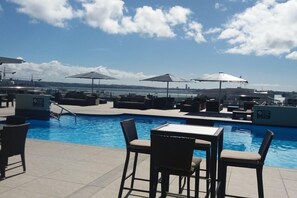 Área de piscina com espreguiçadeiras e guarda-sóis, com vista para um corpo de água e o horizonte distante da cidade.