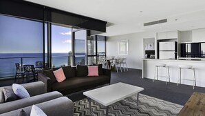 A modern living room with a large window overlooking the ocean, a sofa, a coffee table, and a kitchen area with bar stools. - Circle on Cavill - Private Apartments (Surfers Paradise)