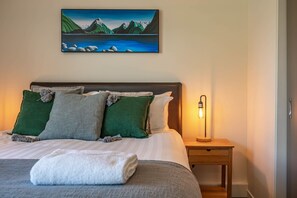Un lit bien fait, recouvert d’une couverture blanche, avec des oreillers verts et gris, et un tableau représentant un paysage montagneux accroché au-dessus.