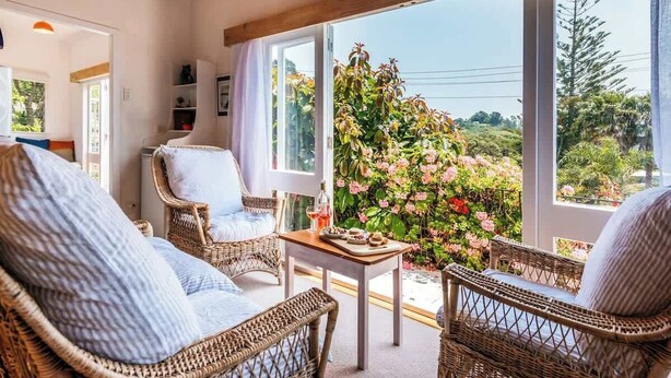 Una habitación acogedora con muebles de mimbre, una mesita y vistas a un jardín lleno de flores rosadas.