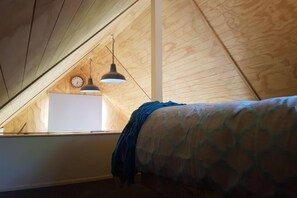 Une chambre avec un plafond en pente, des murs en bois, un lit recouvert d’une couvre-lit à motifs et deux lampes suspendues.