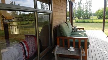 Eine Holzterrasse mit einer Couch und einem Tisch, ein großes Fenster mit Blick auf eine Rasenfläche und ein kleiner Tisch mit einer Tasse.