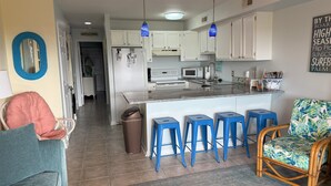 Une cuisine moderne avec des meubles blancs, un îlot central doté de tabourets de bar et un coin repas comprenant une chaise bleue et une chaise en osier.