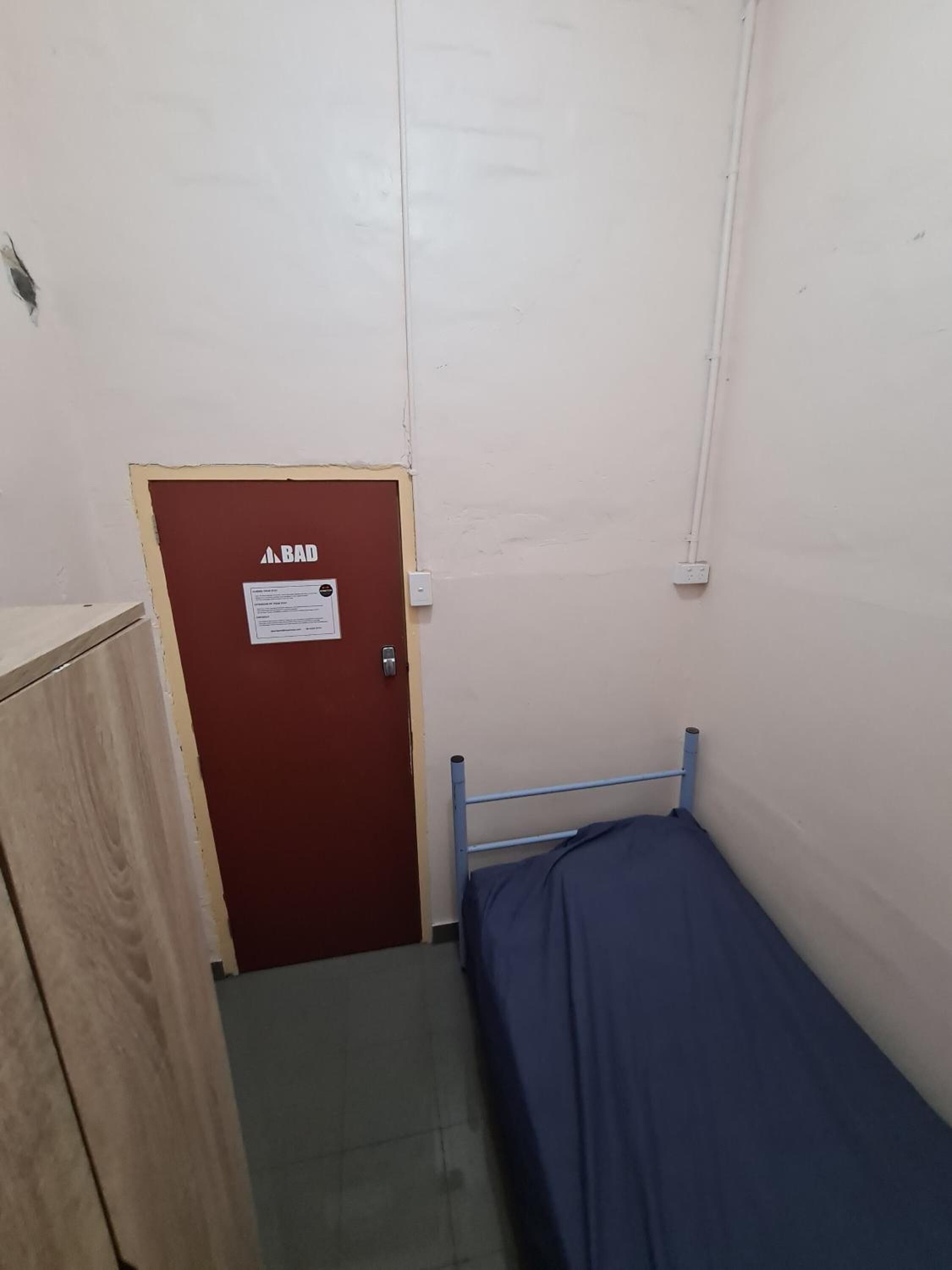 A small, narrow room with a single bed, a wooden cabinet, and a door labeled "BAD".