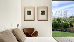 A bedroom with a bed, two framed pictures on the wall, a brown chair, and a view of a garden. - Award Winning Beach House - 100m from Beach (Omaha Beach)