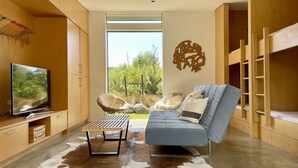 A modern living room with a sofa, a television, a wooden cabinet, and a large window offering a view of greenery. - Award Winning Beach House - 100m from Beach (Omaha Beach)