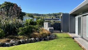 A modern house with a stone retaining wall, a well-maintained lawn, and a view of hills in the background. - Award Winning Beach House - 100m from Beach (Omaha Beach)