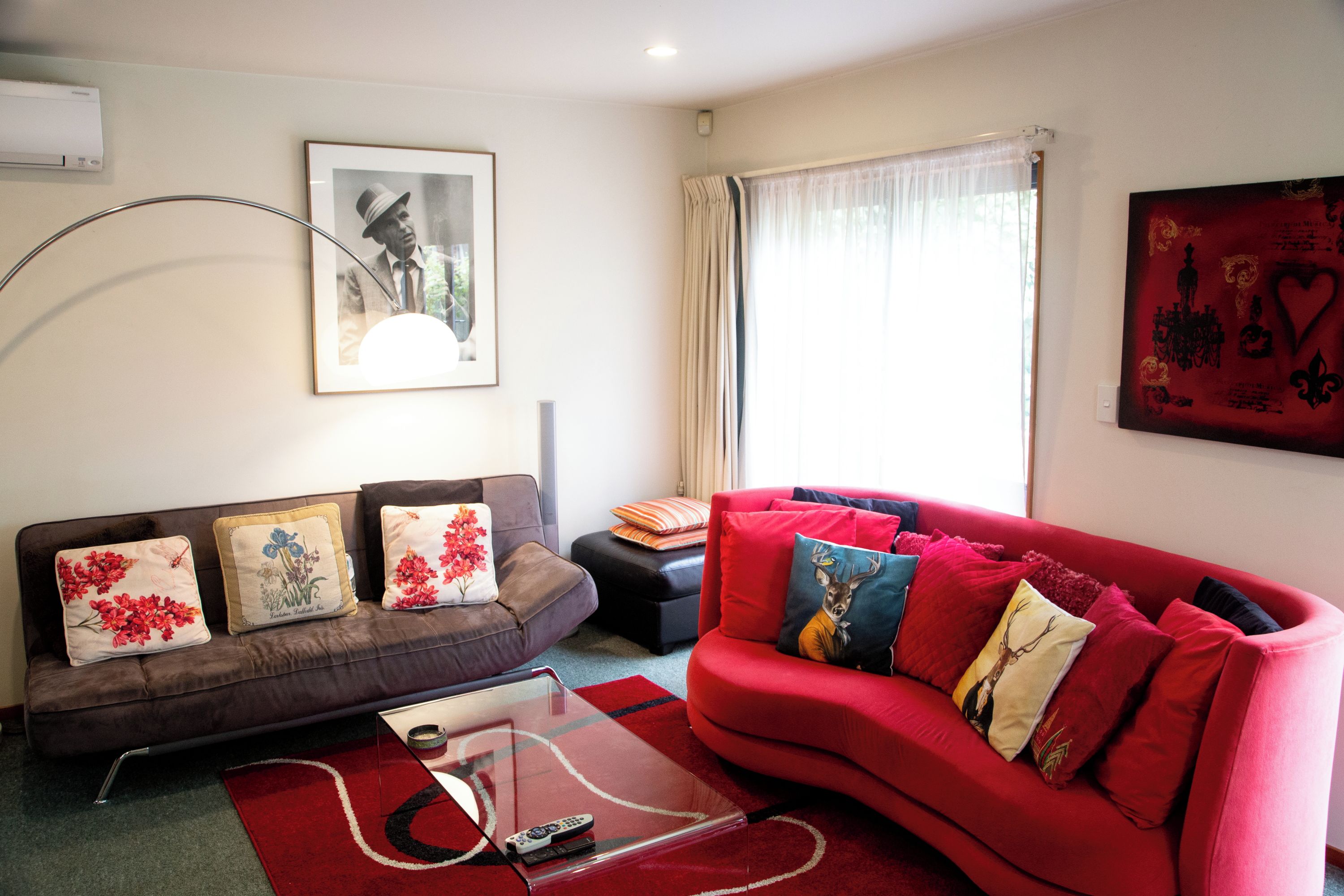 A living room with a brown sofa, red sofa, and a glass coffee table. There is a framed picture on the wall and a window with curtains.