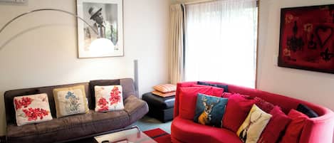 A living room with a brown sofa, red sofa, and a glass coffee table. There is a framed picture on the wall and a window with curtains.
