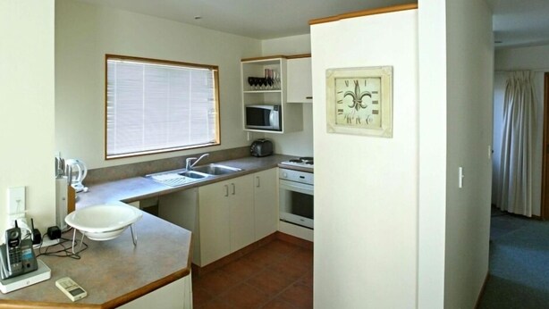 Cocina compacta con fregadero, microondas y estufa. Hay una ventana con persianas, un reloj en la pared y un teléfono en la encimera.