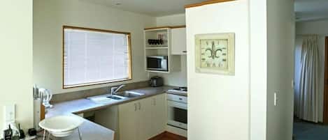 Cocina compacta con fregadero, microondas y estufa. Hay una ventana con persianas, un reloj en la pared y un teléfono en la encimera.